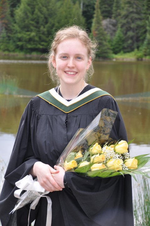 Holly in a convocation robe, holding yellow flowers and standing in front of the campus pond