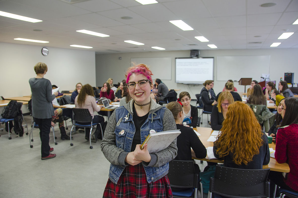 Woman with pink hair stands at the back of a full classroom.