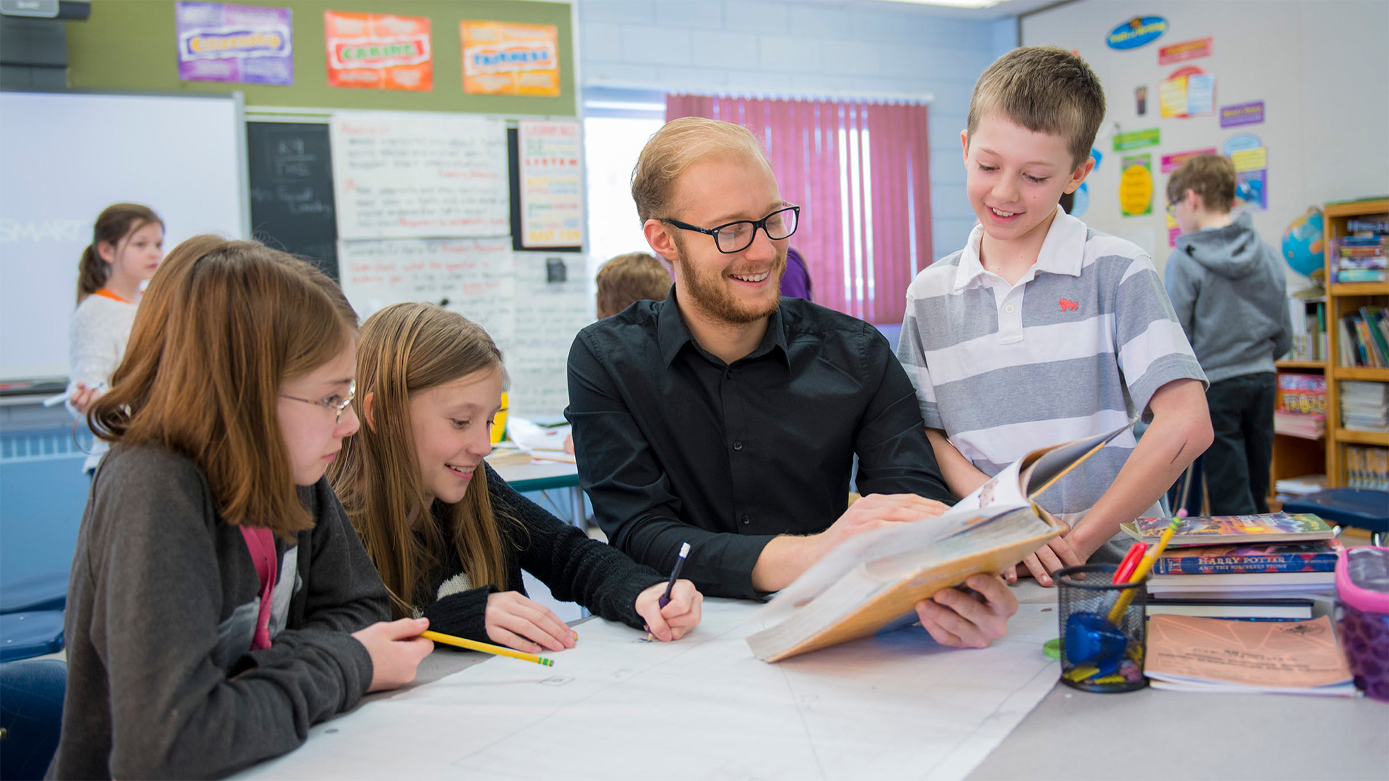A teacher in black shirt reading to a student while some more students sits around them