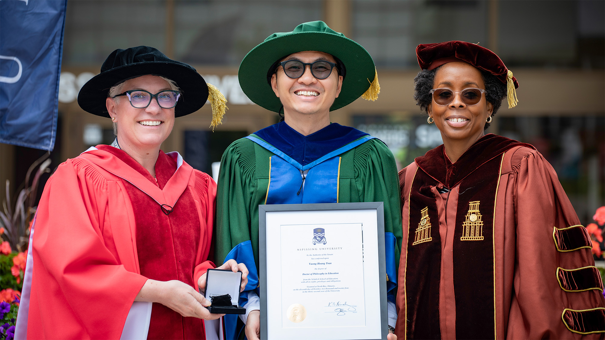 A graduate in green convocation gown holding his degree while 2 professors in red and brown gown stand on either side of him