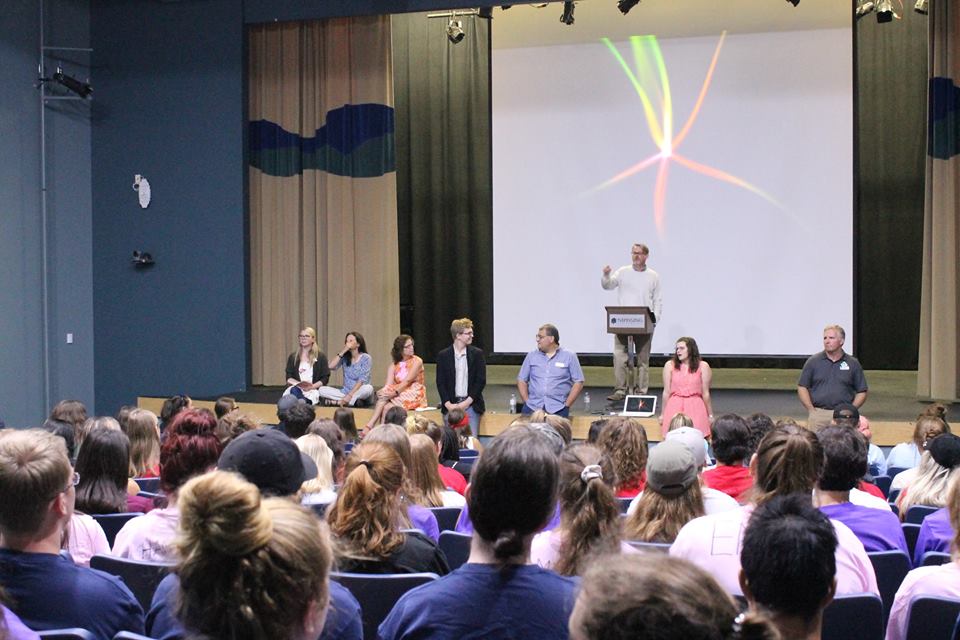 Picture in an auditorium with a group of professors speaking to students
