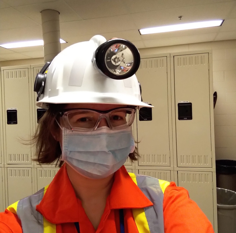 Wearing a bright orange safety suit, white hard hat with light, mask, and safety glasses