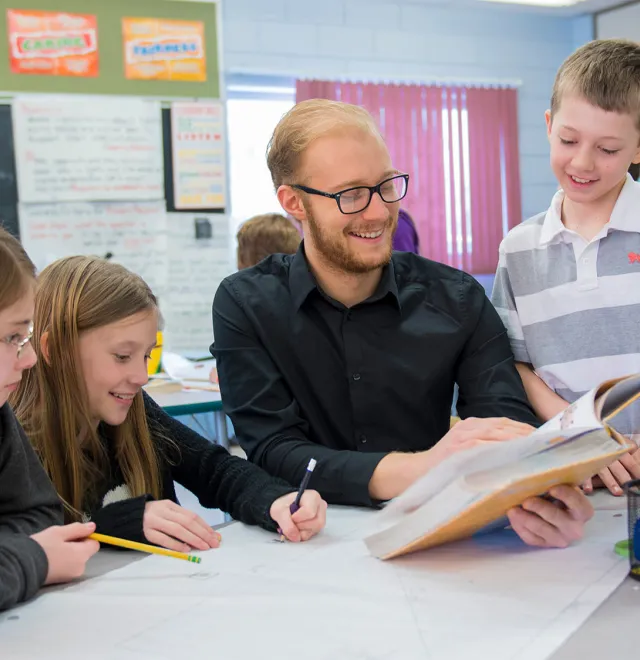 A teacher in black shirt reading to a student while some more students sits around them