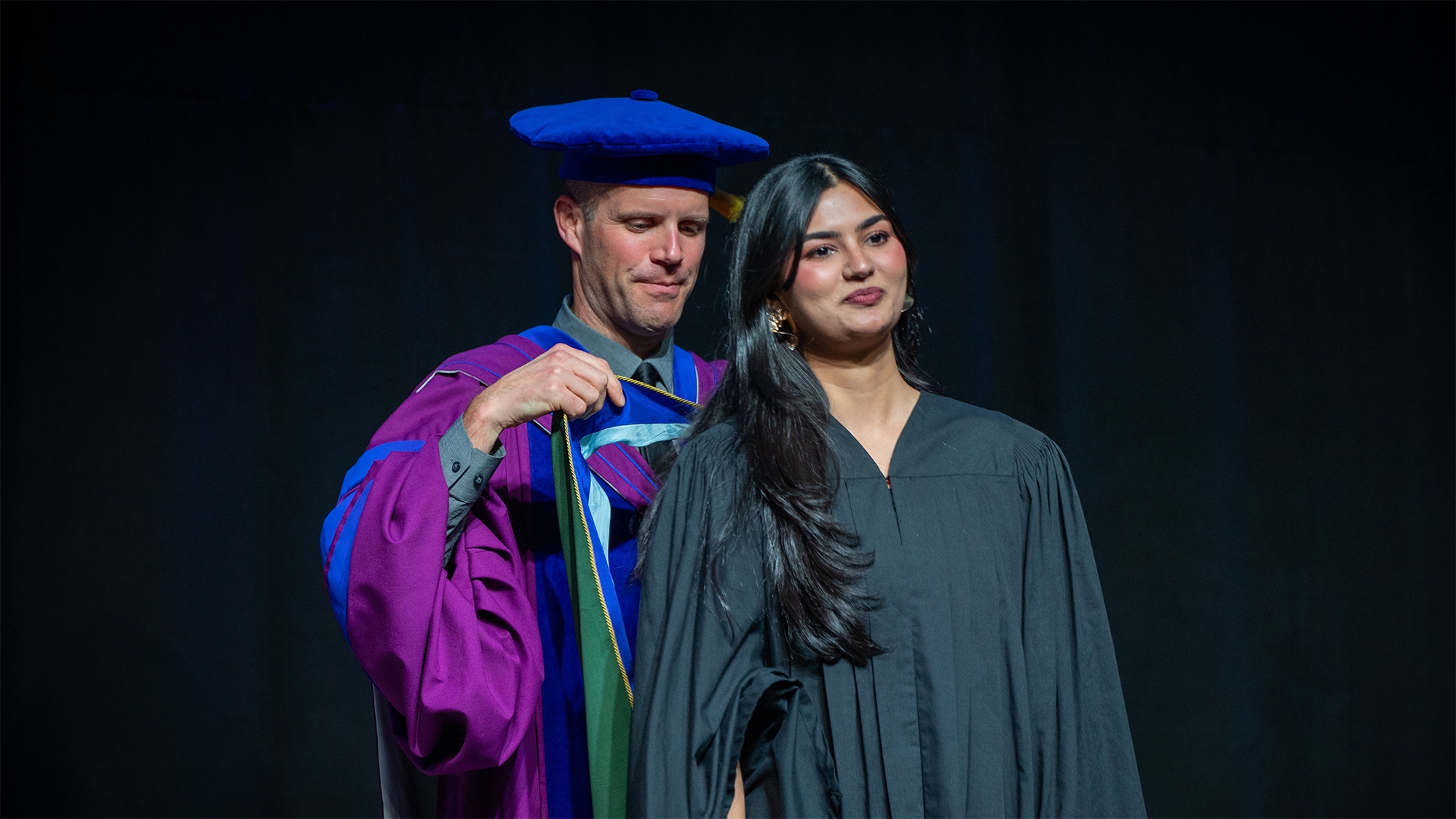 A professor in purple and blue convocation gown putting a ribbon on a graduate in black