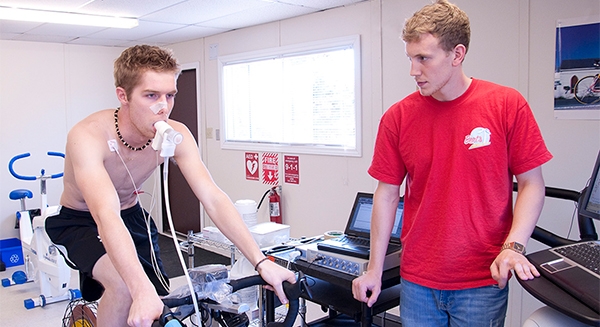 Young shirtless man wearing sensors pedaling a cycle ergometer while a lab technician in a red shirt monitors data