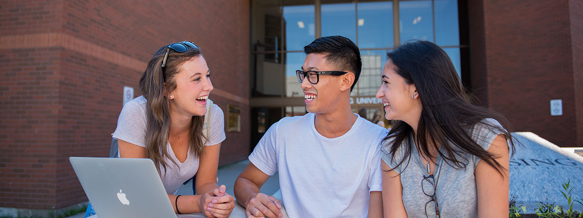 Three students sit on bench outside the front entrance to Nipissing University.