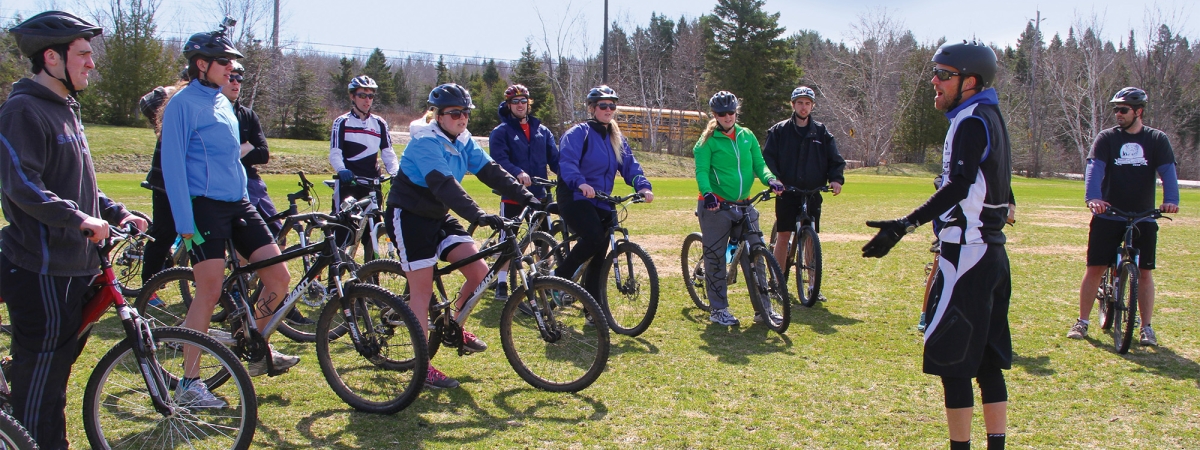 A professor in black outdoor biking gear talking to a group of students on bikes in the field