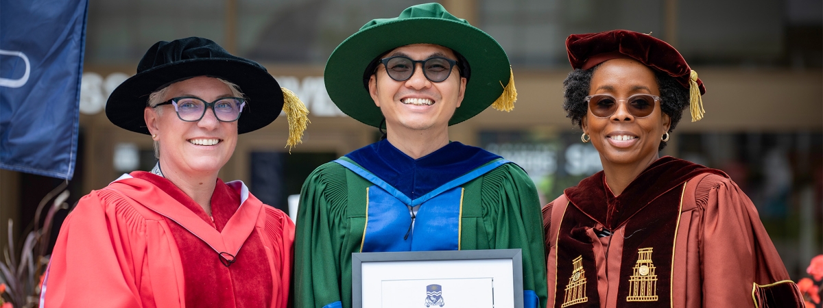 A graduate in green convocation gown holding their degree with 2 professors on both sides of him