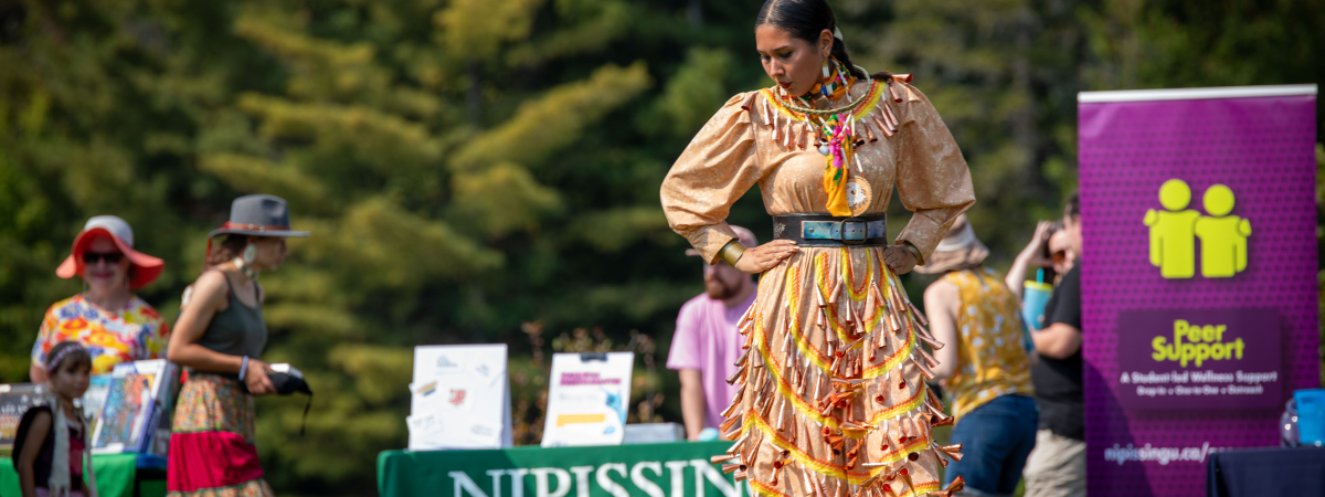 Pow wow dancer in yellow regalia beside the campus pond.