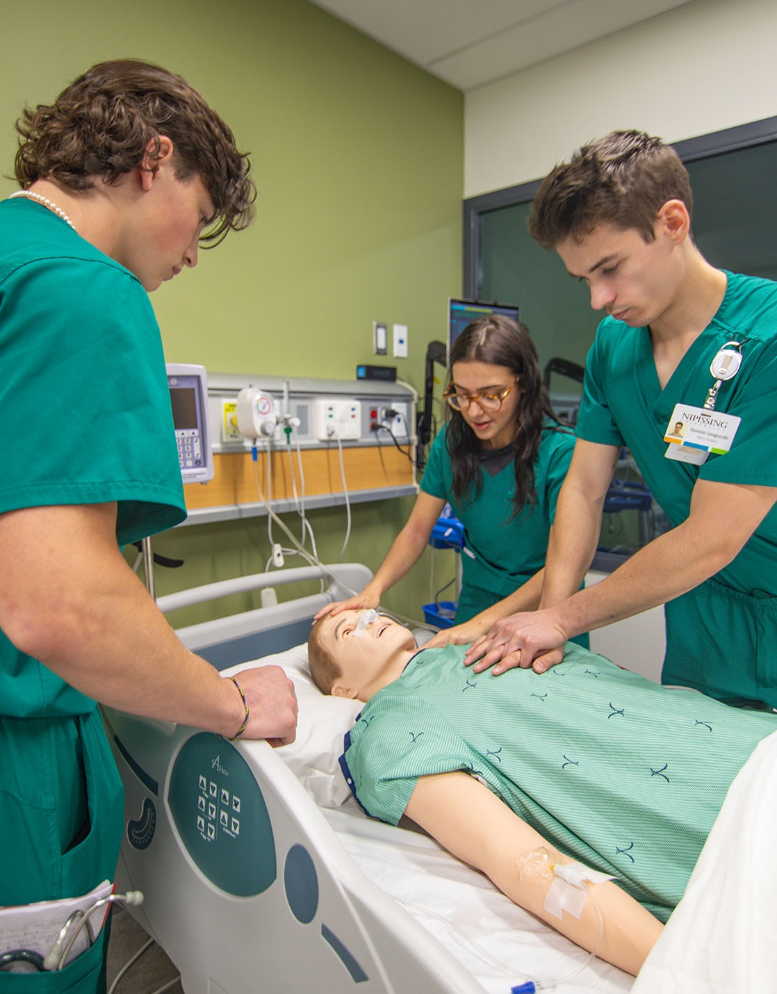 3 students in green scrubs doing chest compression on a mannequin in the simulation lab at Nipissing